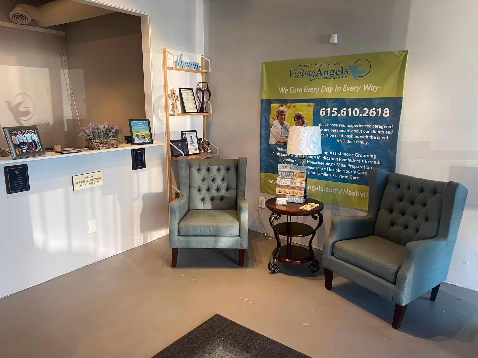 Two teal armchairs sit beside a round table under a green Visiting Angels banner.