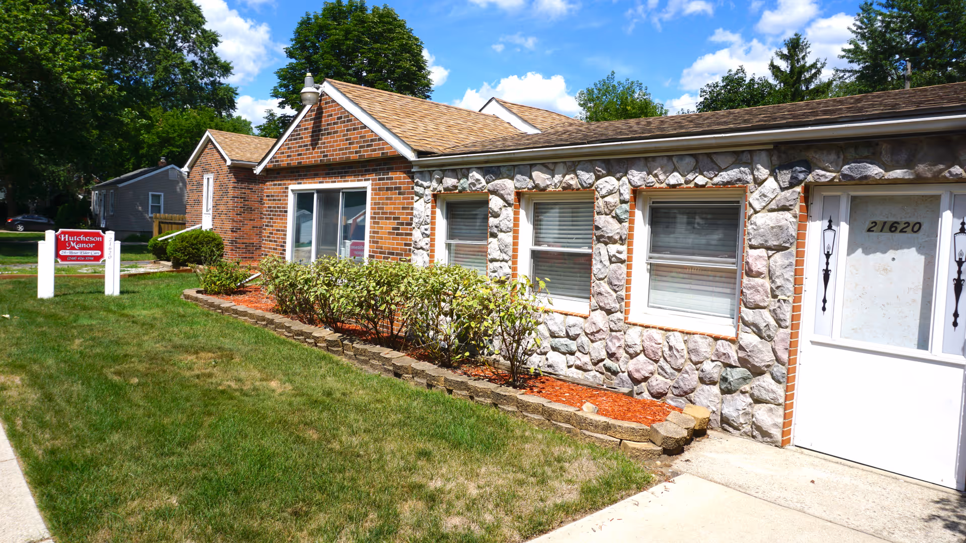 Exterior view of Hutcheson Manor Nursing Home Alternative building with stone and brick facade, three windows, a white door with the number 21620, and a green lawn with bushes in front. A red and white sign with the facility name is visible on the lawn.