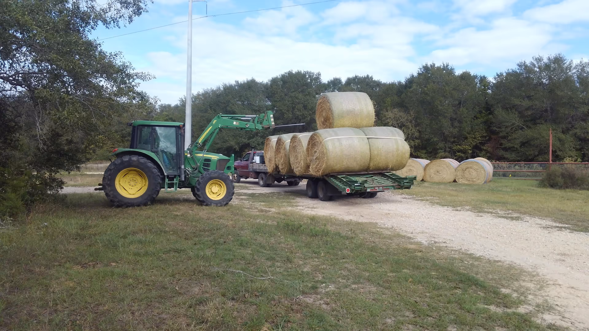 Green tractor with hay bale trailer on dirt road beside stacked round bales.