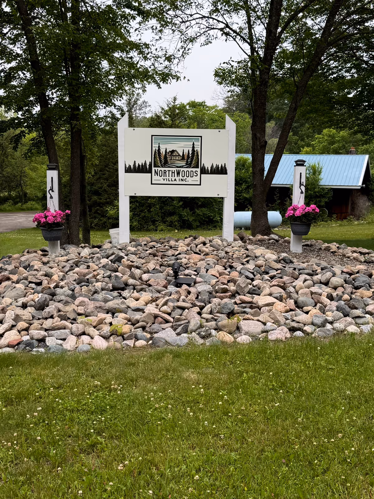 An entrance sign reading "Northwoods Villa Inc." on white posts surrounded by a rock bed, trees, and a building with a blue roof in the background.