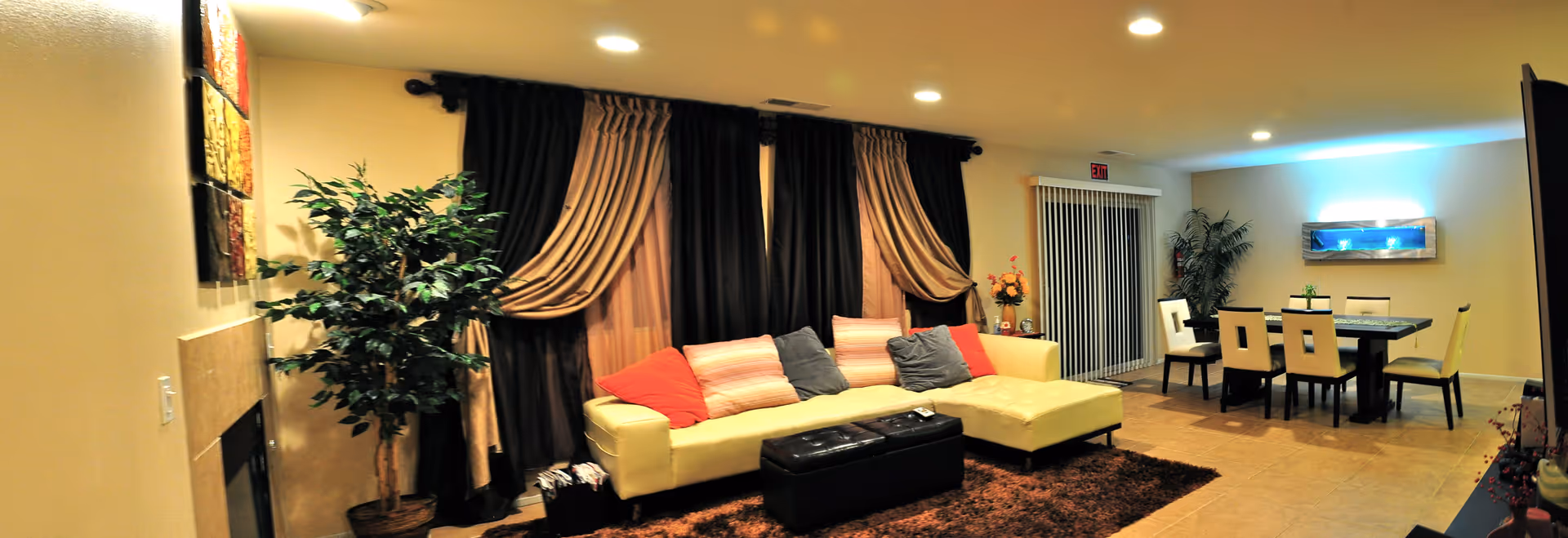 Open living and dining area with a cream sectional sofa, dark drapes, a black ottoman, and a dining table in the background.