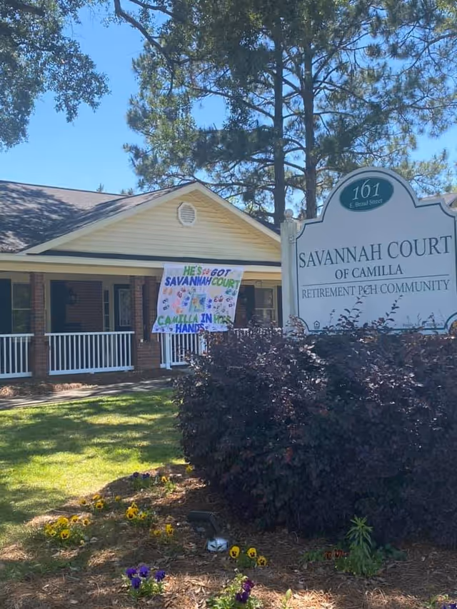 Exterior view of Savannah Court of Camilla retirement community building with a white sign displaying the facility name and address. The building has a porch with white railings and a banner hanging that reads 'He's got Savannah Court Camilla in his hands.' There are trees and flower beds with yellow and purple flowers in the foreground.