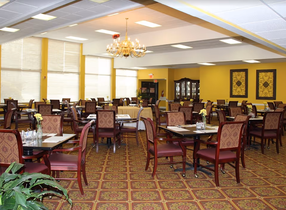 Spacious dining room with multiple set tables and upholstered chairs, chandeliers, yellow walls and patterned carpet.