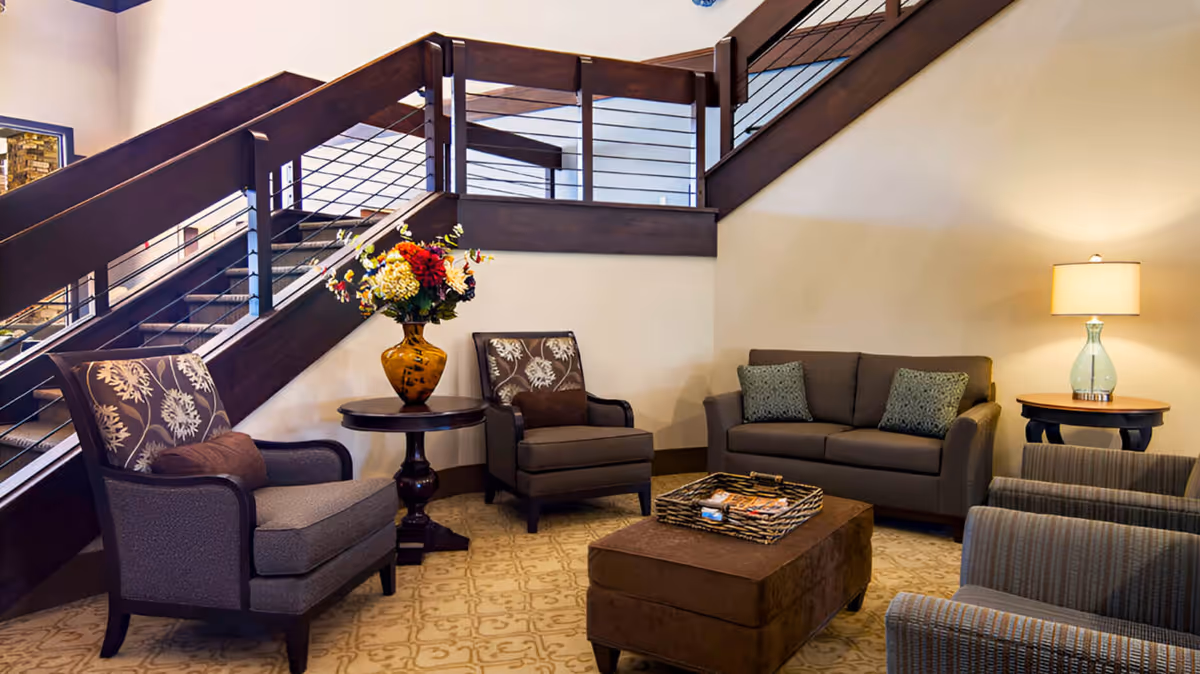 Comfortable lobby seating area with armchairs, a sofa, ottoman, side tables, a lamp, floral arrangement, and a staircase in the background.