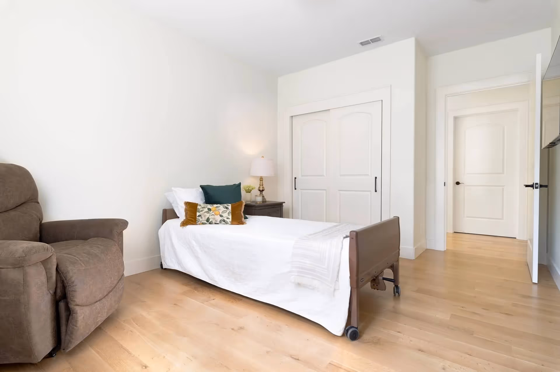 Bright bedroom with a hospital-style adjustable bed, recliner, and nightstand in a senior living room.