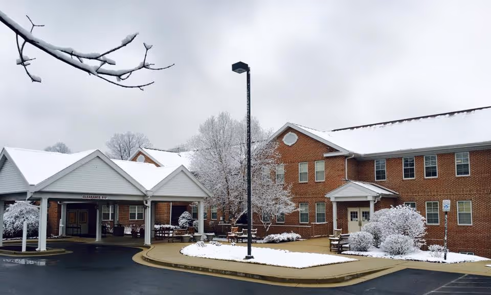 Exterior view of Sterling Care Hillhaven facility in winter with snow covering the roof, trees, bushes, and ground. The building is made of red brick with white trim and has a covered entrance with a clearance sign. A streetlamp stands in front of the building along a curved driveway.
