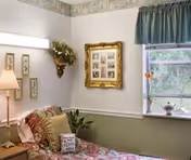 Cozy bedroom with a floral bedspread, framed pictures on the wall, a lamp, decorative plants, and a window with a green valance.