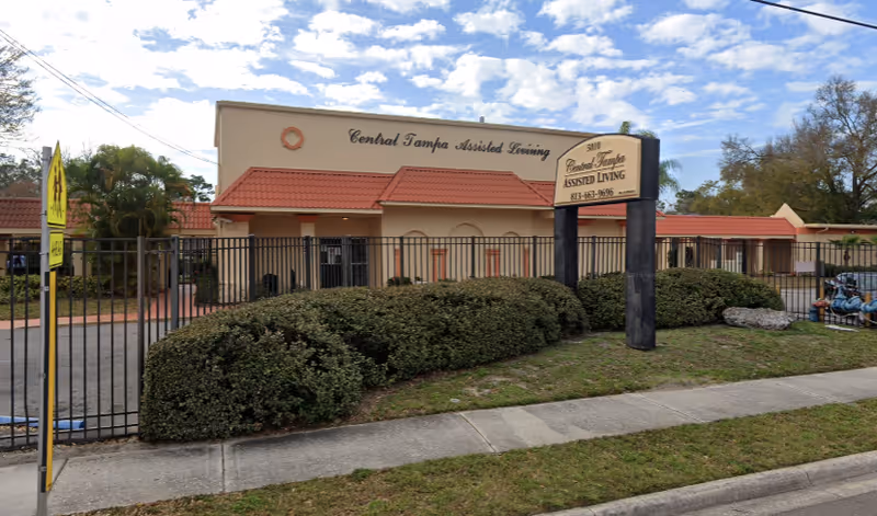 Exterior view of Central Tampa Assisted Living facility showing a beige building with a red tiled roof, a black metal fence, bushes in front, a sidewalk, and a sign with the facility name and phone number.
