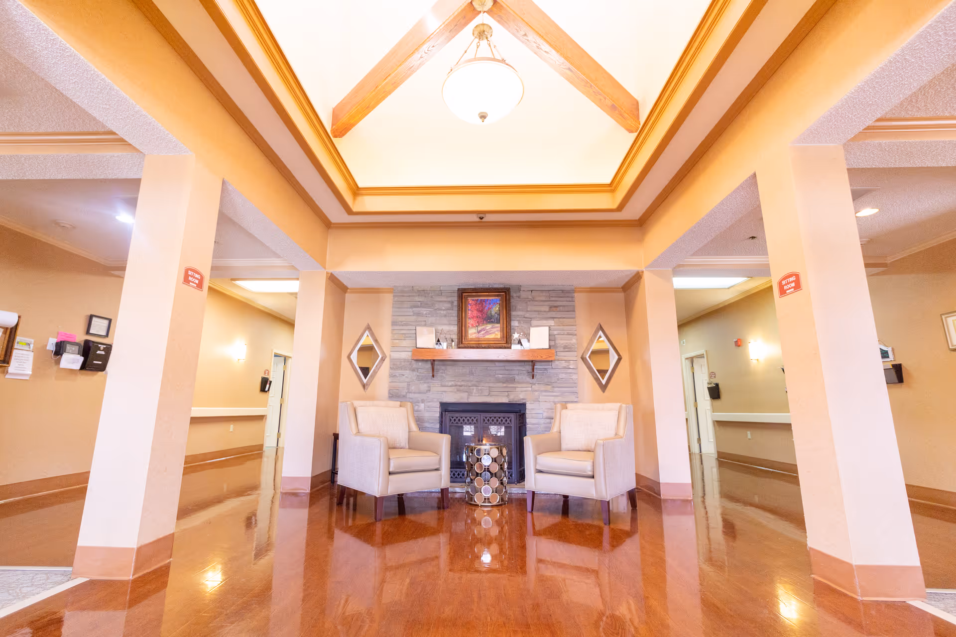 A bright and spacious common area with two beige armchairs facing each other in front of a stone fireplace. Above the fireplace is a colorful framed painting, and two diamond-shaped mirrors are mounted on the wall on either side. The room features a high ceiling with wooden beams and a hanging light fixture. The floor is polished wood, and there are hallways visible on both sides of the seating area.