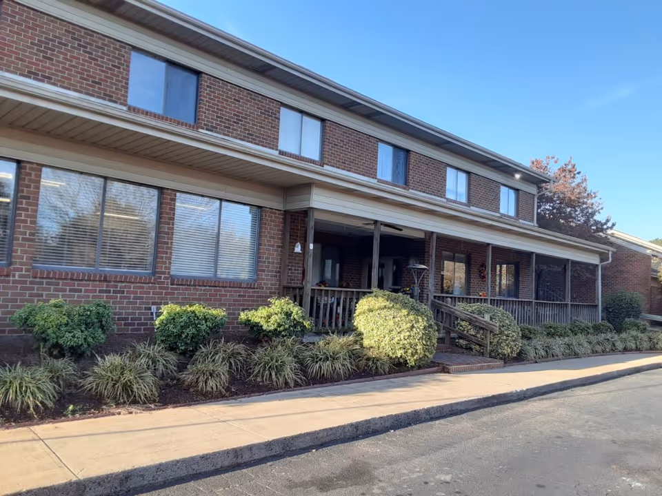 Two-story brick building with a covered front porch, ramp, and landscaped shrubs under a clear sky.