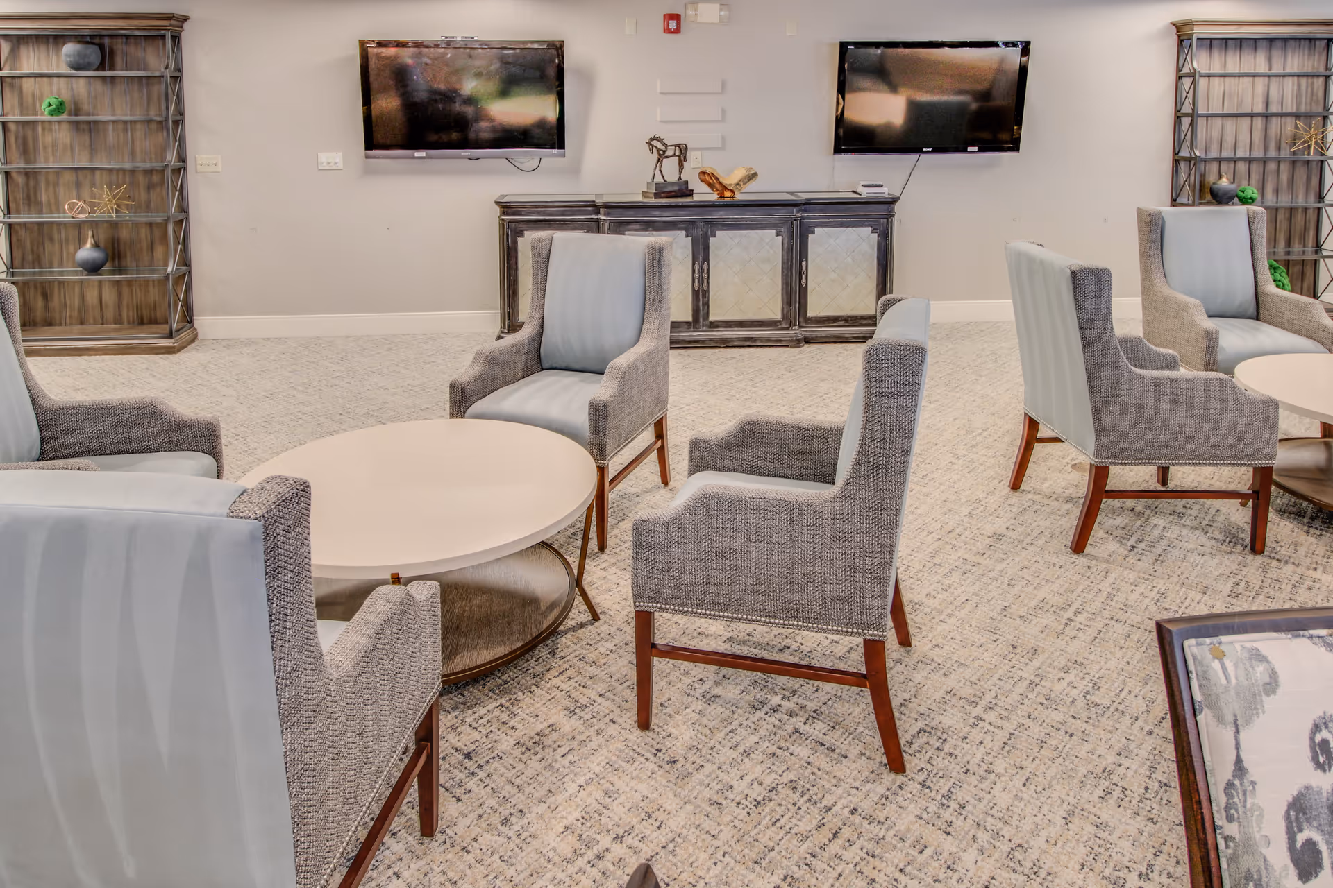 Cozy senior living lounge with round tables, gray upholstered chairs, and wall-mounted TVs.