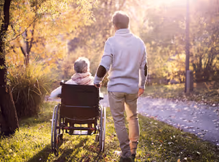 Autumnal park path with an elderly person in a wheelchair and a caregiver.