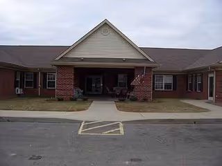 Front view of a brick senior living facility entrance with a small porch and chairs.