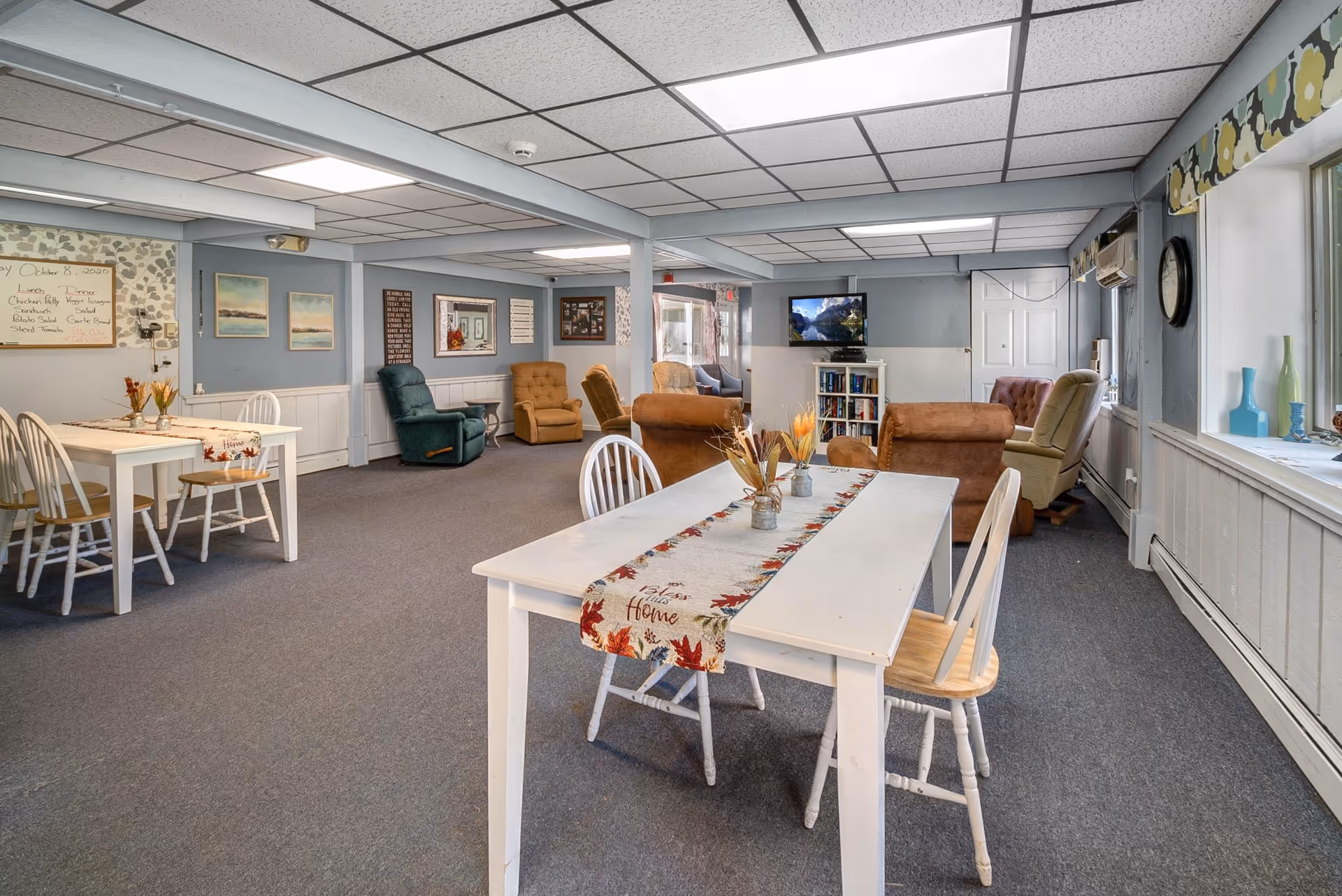 Communal senior living dining and lounge area with white tables and chairs, recliner armchairs, and a television.
