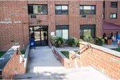Exterior brick senior living center entrance with stairs, ramp, planters, and people nearby.
