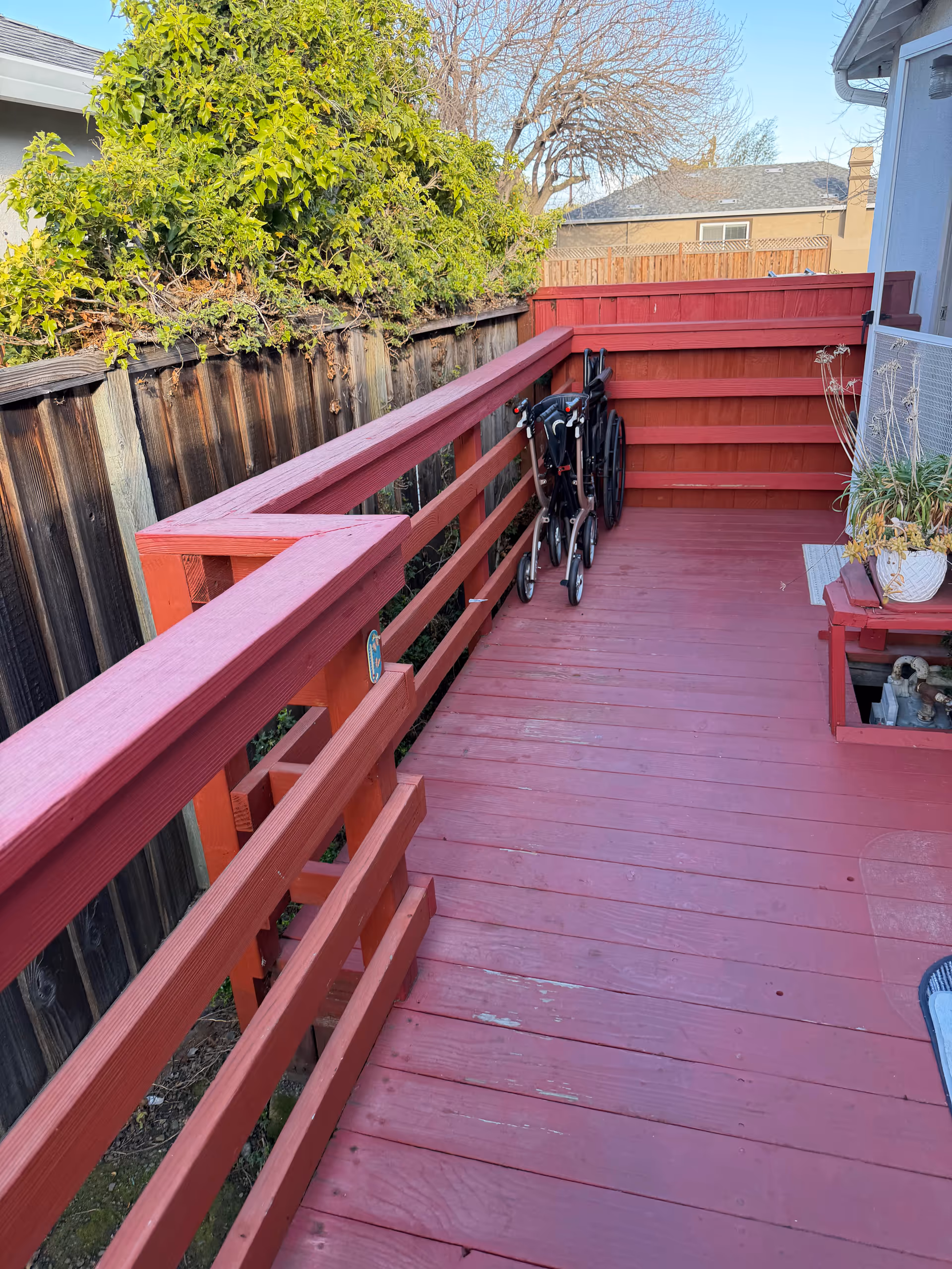 Red wooden outdoor deck with railing, wheelchairs by wall and potted plant near doorway.