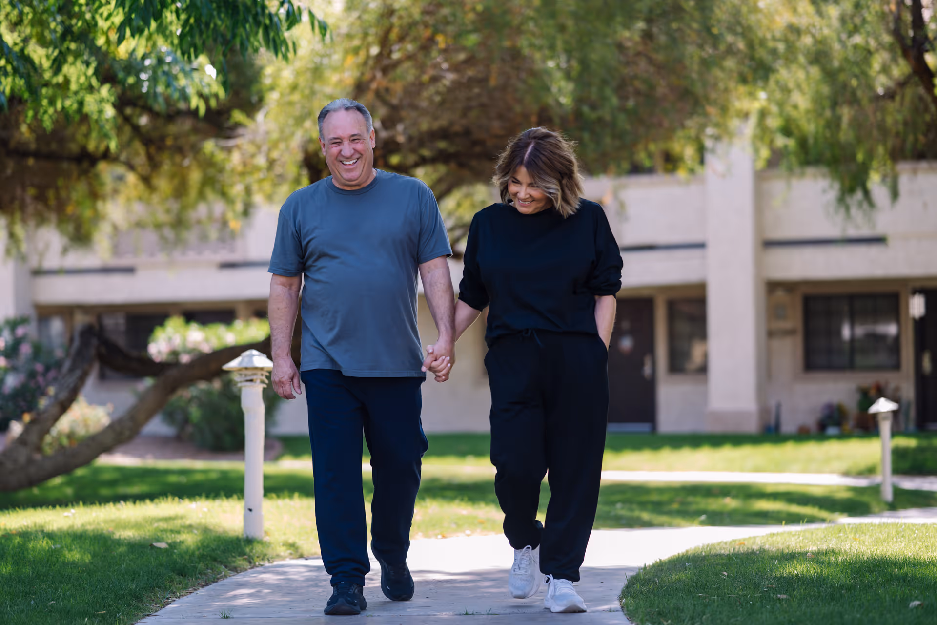 An older man and woman holding hands and walking on a paved path in a green outdoor area with trees and a building in the background, both smiling and dressed casually.