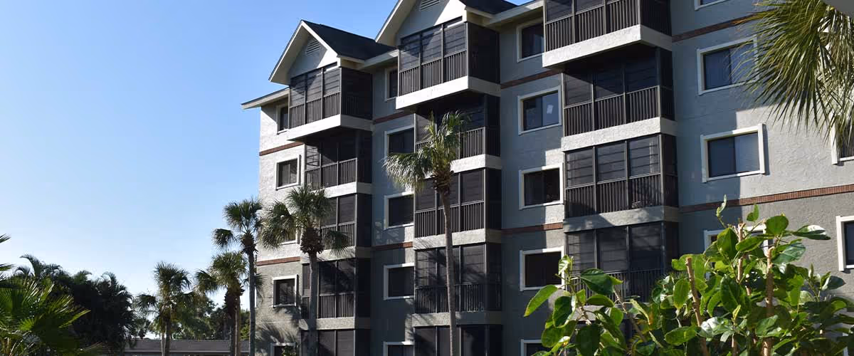 Exterior view of a multi-story apartment building with screened balconies and palm trees under a clear blue sky.