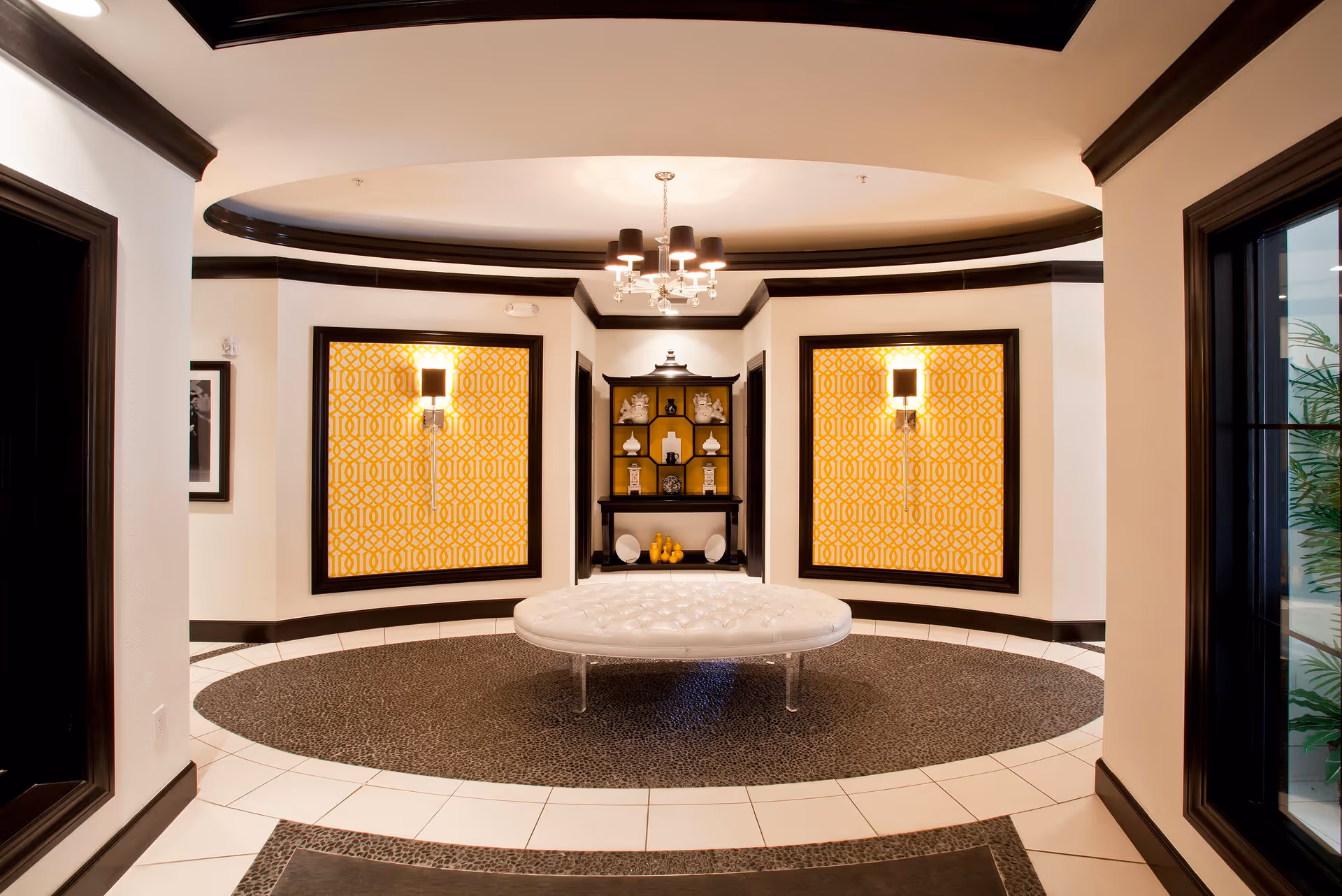 A stylish circular lobby area with a white tufted round bench in the center, surrounded by a patterned floor. The walls feature two large framed yellow and white geometric patterned panels with wall sconces above them. A dark wood display shelf with decorative items is centered between the panels. The ceiling has a recessed circular design with a chandelier hanging in the middle. The space is accented with dark trim and white walls.
