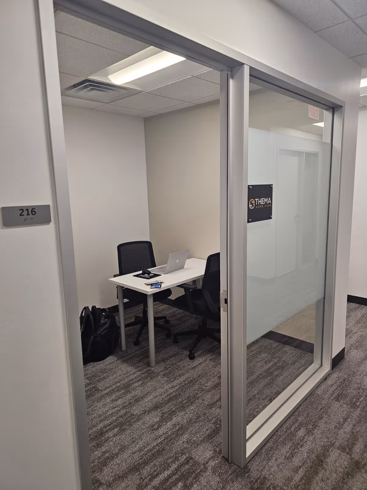 Small glass-walled office with desk, laptop, and chairs inside Thema Home Care facility.