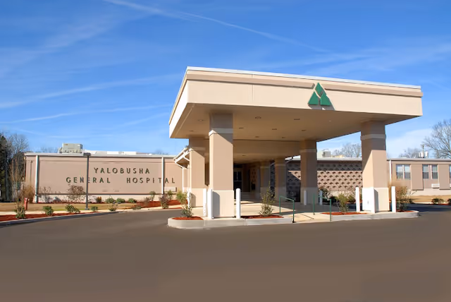 Exterior view of Yalobusha General Hospital & Nursing Home building with a covered entrance supported by columns, clear blue sky, and landscaped area with small plants and bushes.