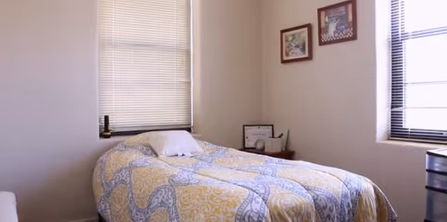 A neatly made single bed in a small bedroom with two windows and framed art.