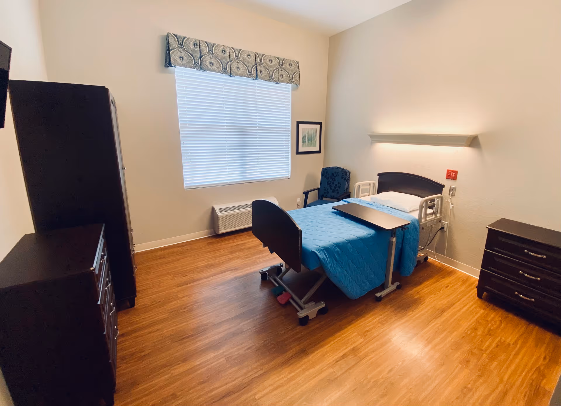 A furnished senior living bedroom with a blue hospital bed, dresser, chair, and window.