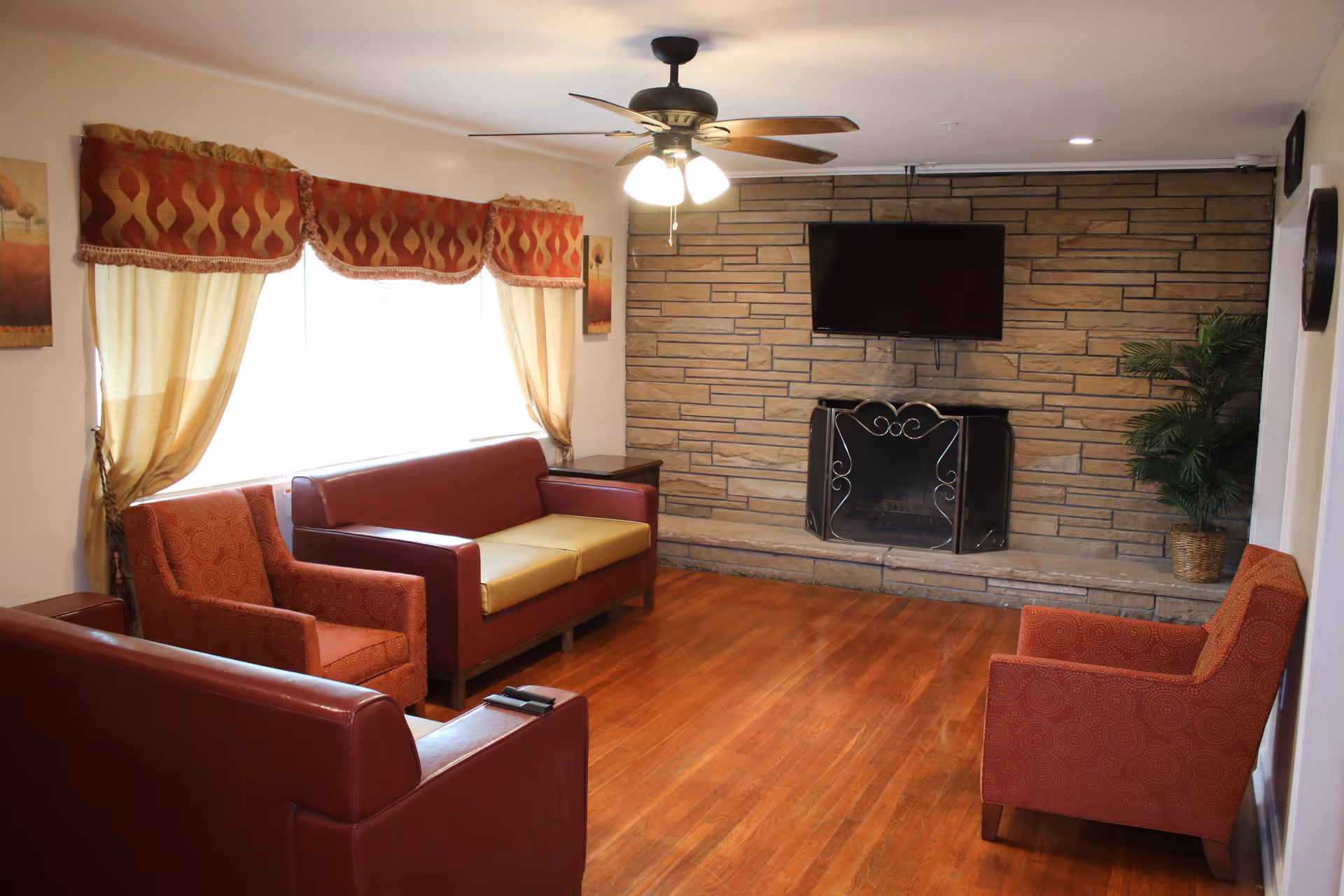 A cozy living room with wooden flooring, featuring two red sofas and two red armchairs arranged around a stone fireplace with a mounted flat-screen TV above it. The room has a ceiling fan with lights, large windows with cream-colored curtains and red valances, and framed artwork on the walls. A potted plant is placed near the fireplace.