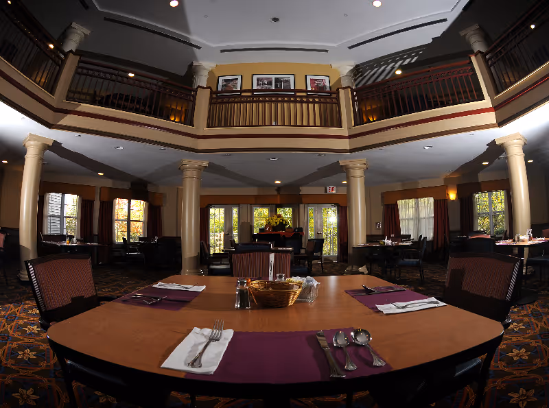 Spacious senior living dining room with set tables, columns, a mezzanine balcony, and large windows.
