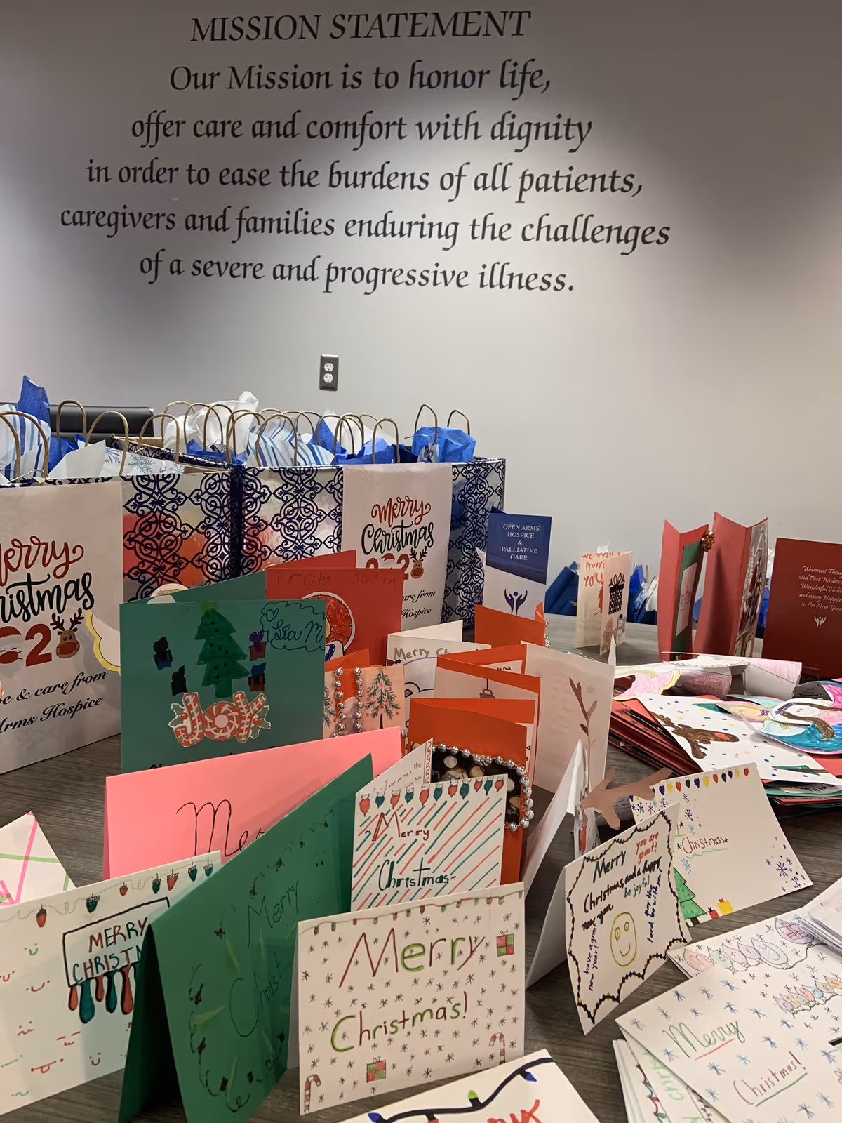 Rows of handmade Christmas cards and gift bags on a table beneath a mission statement wall.