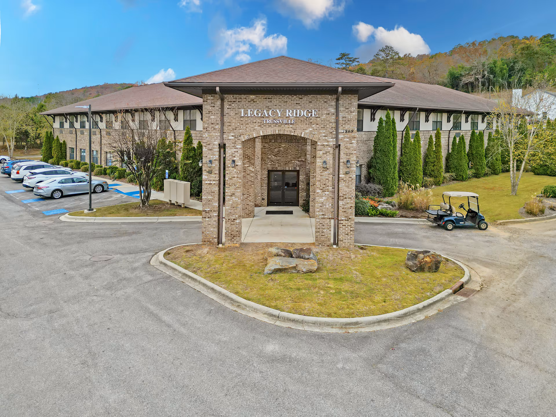 Front exterior view of the Legacy Ridge Trussville building, a two-story brick structure with a covered entrance. There are several cars parked in the parking lot to the left and a golf cart parked on the right side near landscaped greenery and trees. The sky is clear with some clouds.