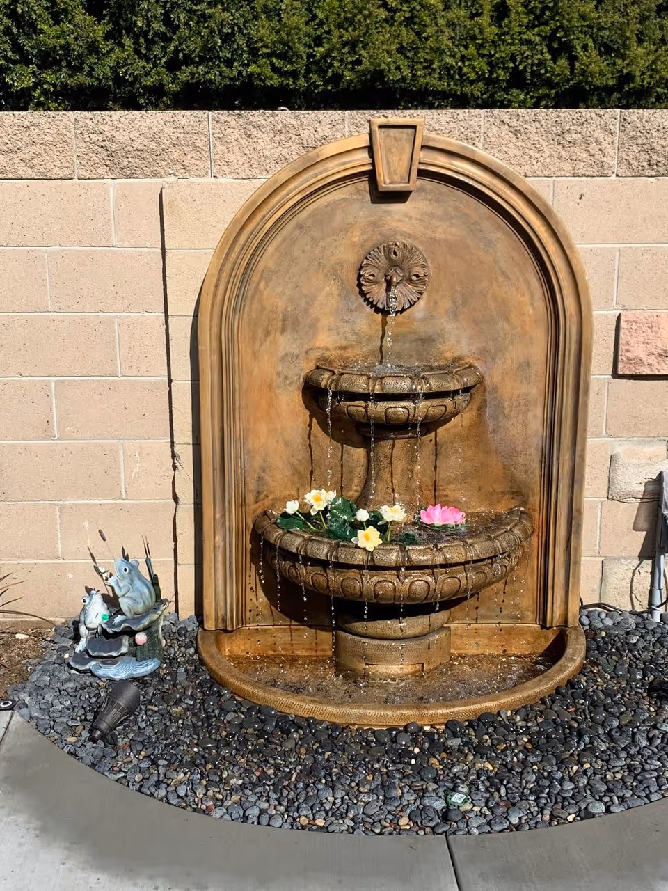 Outdoor wall-mounted two-tier water fountain with water flowing from a decorative spout into the upper basin and then into the larger lower basin, which contains floating artificial flowers. The fountain is set against a beige brick wall with green shrubbery above it. There are decorative frog statues on the left side of the fountain, placed on a bed of small black and gray pebbles.