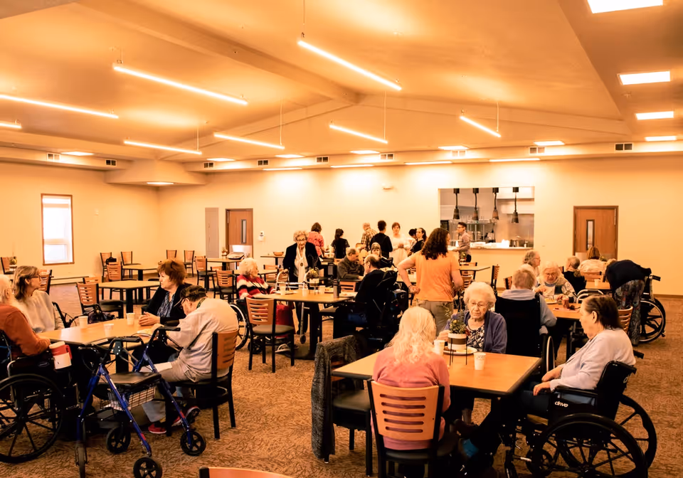 A large dining room in an assisted living facility with elderly residents sitting at tables, some in wheelchairs, engaging in conversation. The room has warm lighting, beige walls, and a kitchen area visible in the background.
