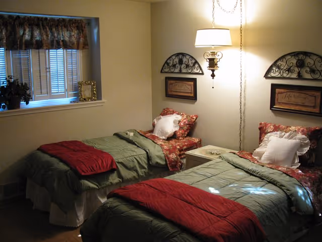 A cozy bedroom with two twin beds, each made with green bedspreads and red blankets at the foot. Both beds have floral-patterned pillows and white decorative pillows. A small nightstand with a lamp is positioned between the beds. The walls are light-colored with decorative wall hangings above each bed. A window with closed blinds and a floral valance is on the left side of the room.