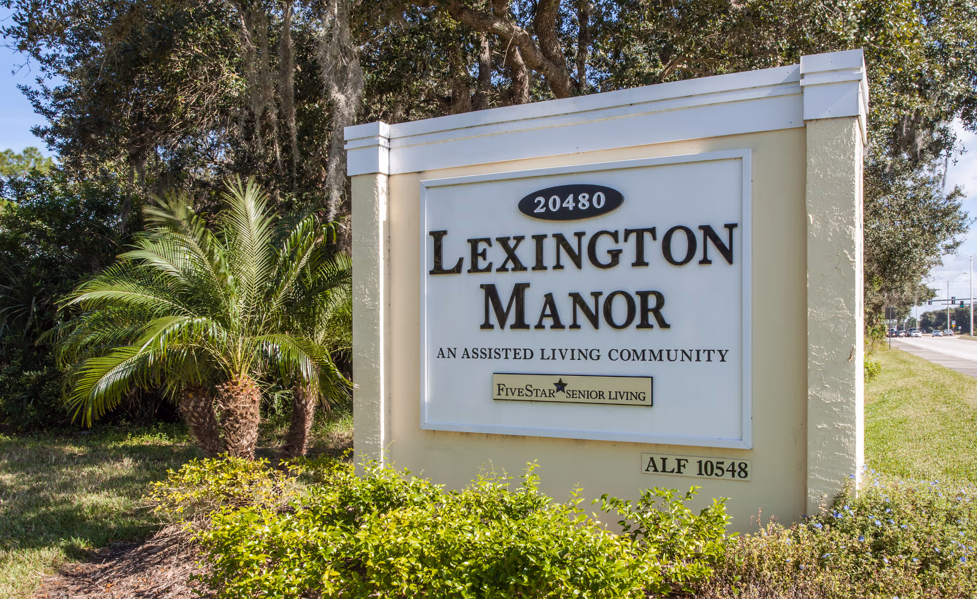 Entrance sign reading "Lexington Manor — An Assisted Living Community" surrounded by landscaping and a nearby road.