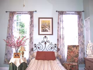 A bedroom scene with a metal headboard bed, two windows, and floral decor.