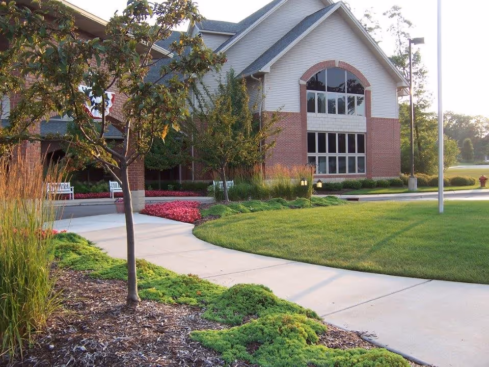 Exterior view of a senior living facility building with a brick and siding facade, large windows, a paved walkway, green lawn, trees, and landscaped flower beds.