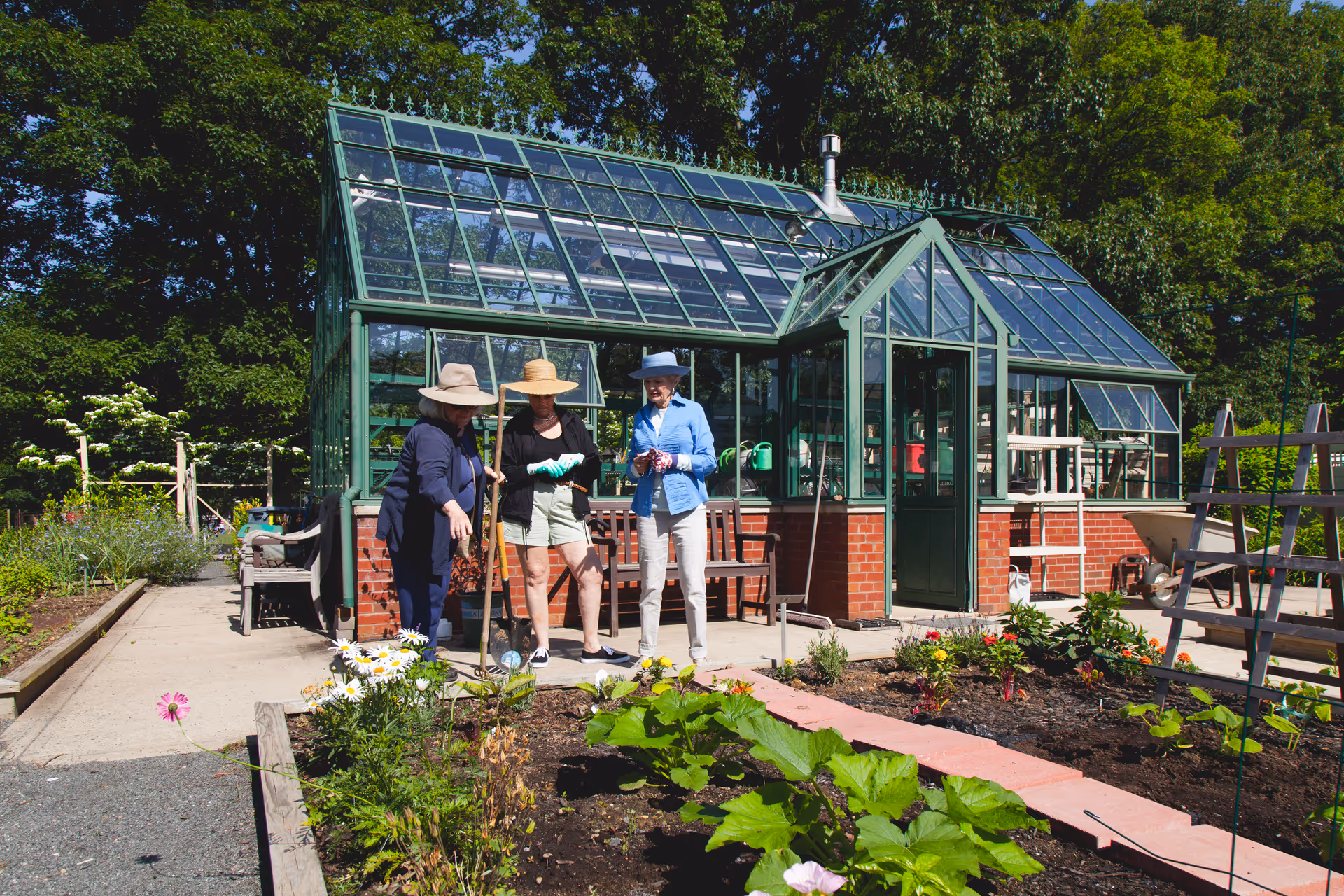 Three people in hats stand near a glass greenhouse in a garden outside.