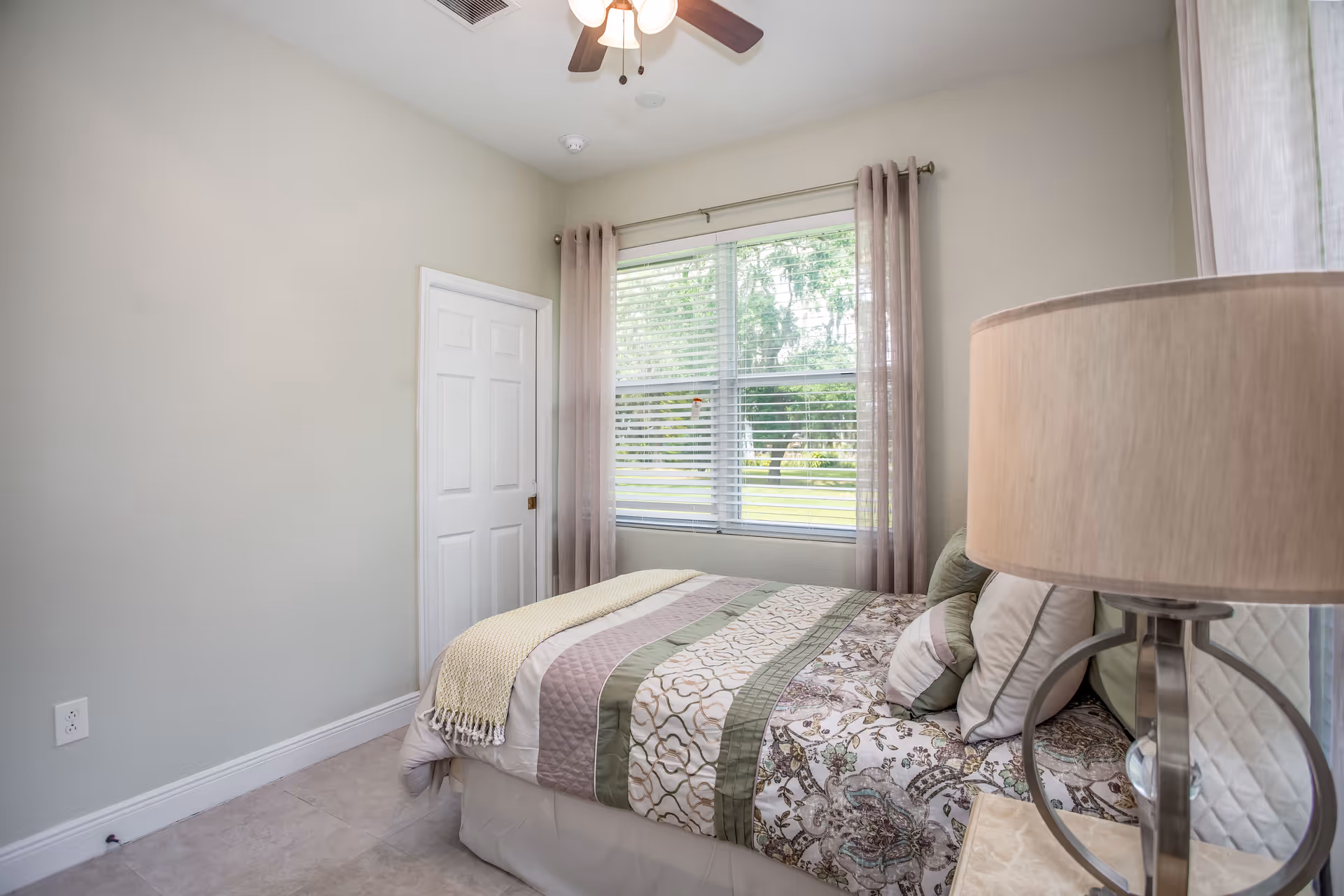 A small bedroom with a bed covered in a patterned quilt and multiple pillows. There is a window with blinds and sheer curtains letting in natural light, a white door, a ceiling fan with lights, and a bedside table with a lamp.