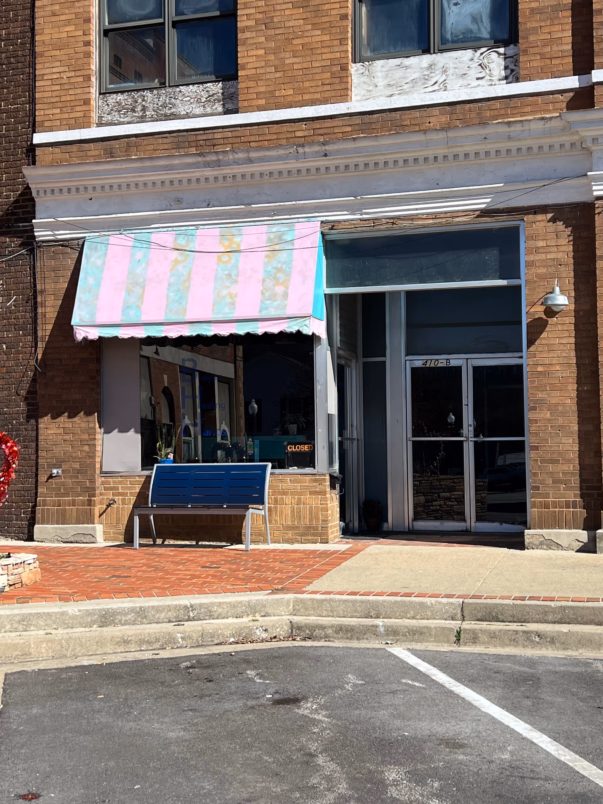 A brick storefront features a pink striped awning and a blue bench outside.