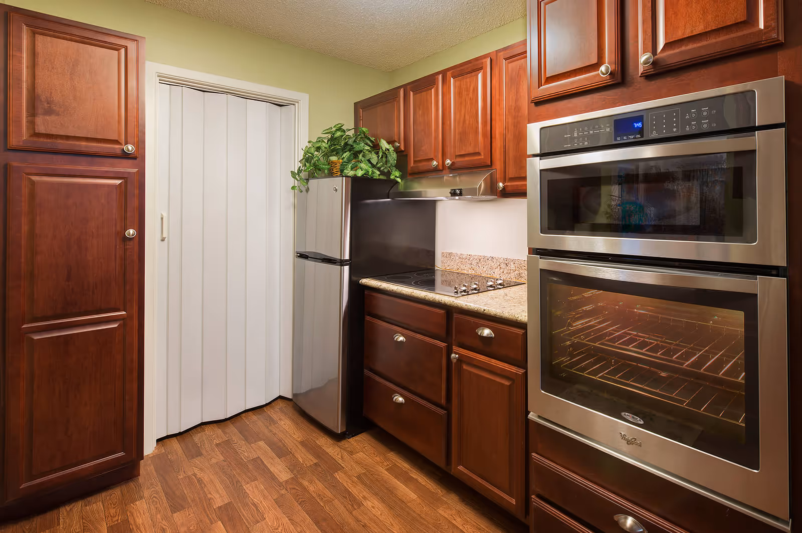 Small kitchen with cherry wood cabinets, stainless steel double oven, compact refrigerator, electric cooktop, and wood-look flooring.