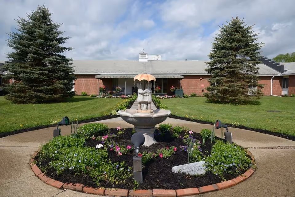 Circular flowerbed with a decorative fountain sculpture centered in front of a single-story brick building and flanked by evergreen trees.