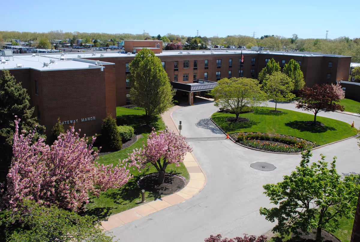 Aerial view of a senior living facility named Gateway Manor with a circular driveway, landscaped green lawns, and blooming trees with pink and green foliage under a clear blue sky.