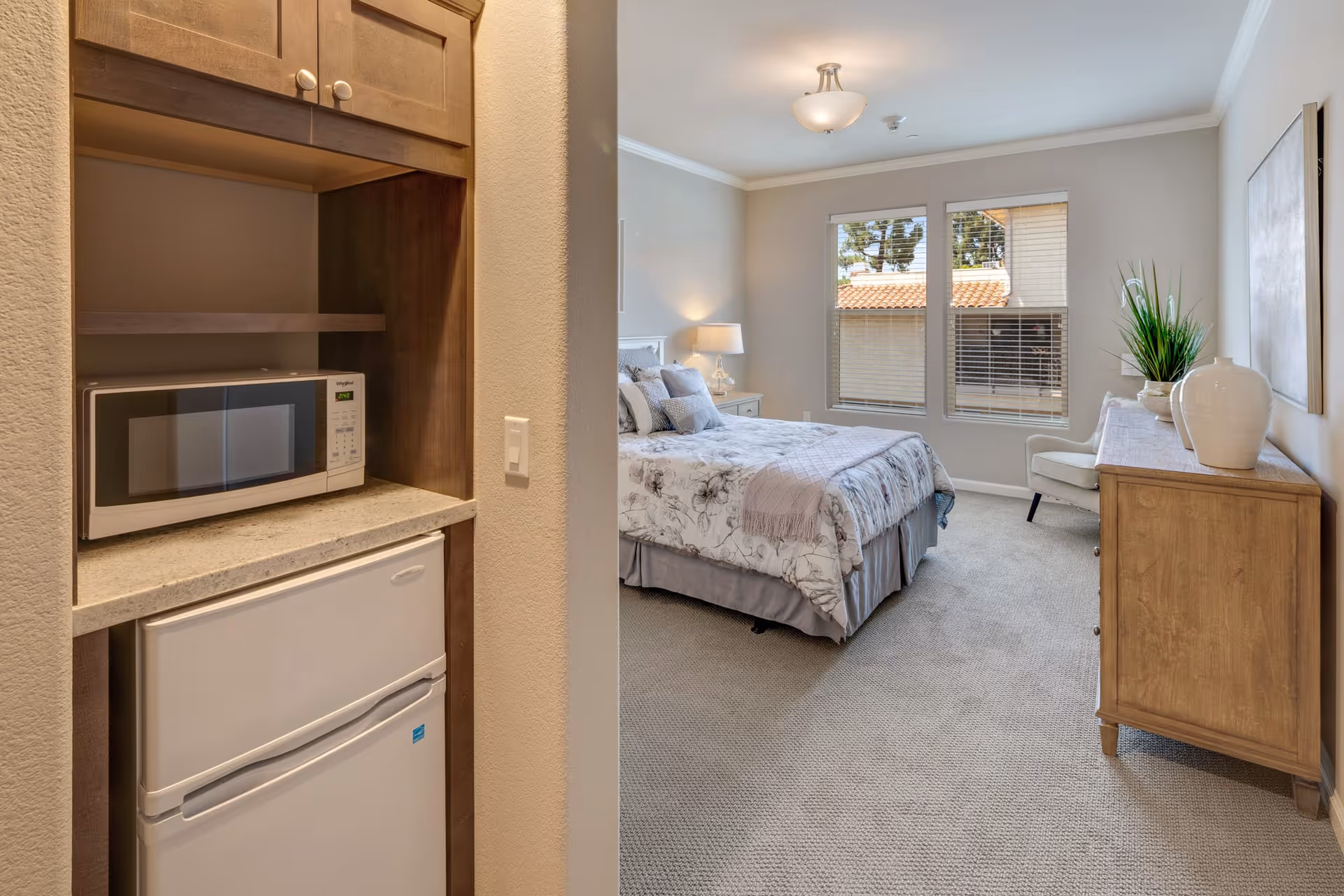 A cozy bedroom with a bed covered in floral bedding, a nightstand with a lamp, a wooden dresser with decorative vases and a plant, and a chair near two large windows letting in natural light. In the foreground, there is a small kitchenette area with a microwave and a mini refrigerator.