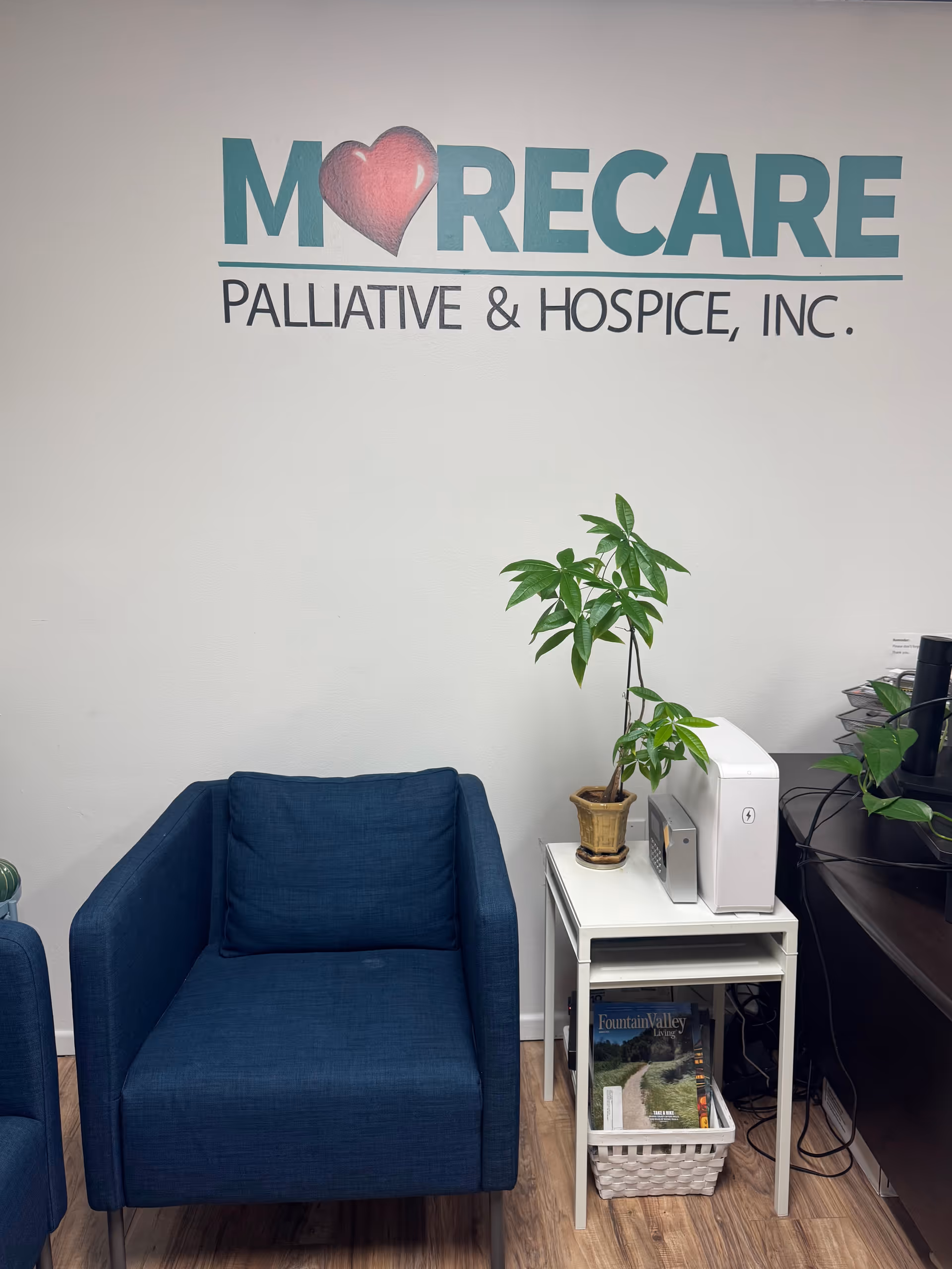 Blue chairs by a white table with a plant, under Morecare Palliative & Hospice logo.