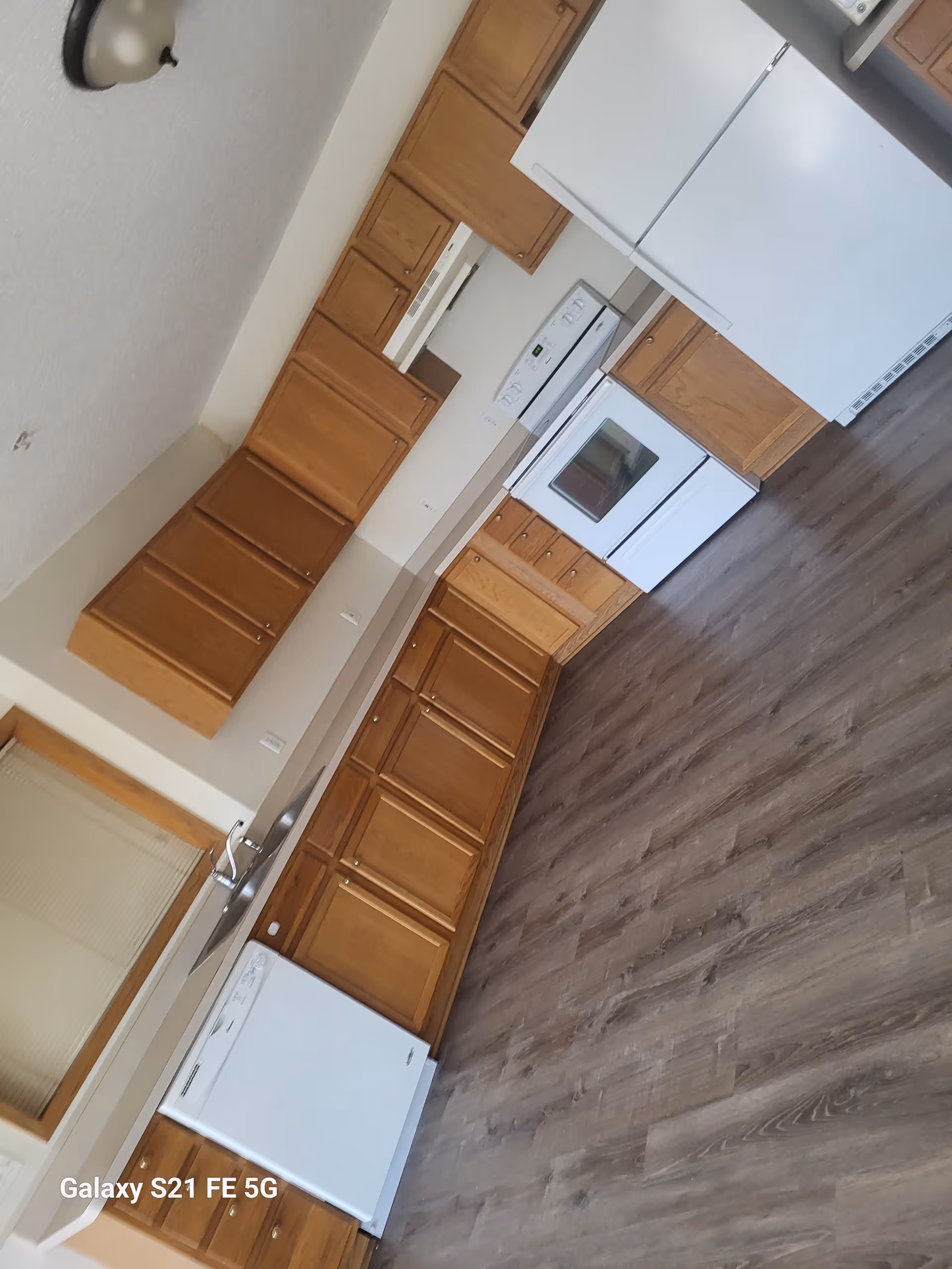 Long kitchen with wooden cabinets, white appliances, and dark wood flooring.