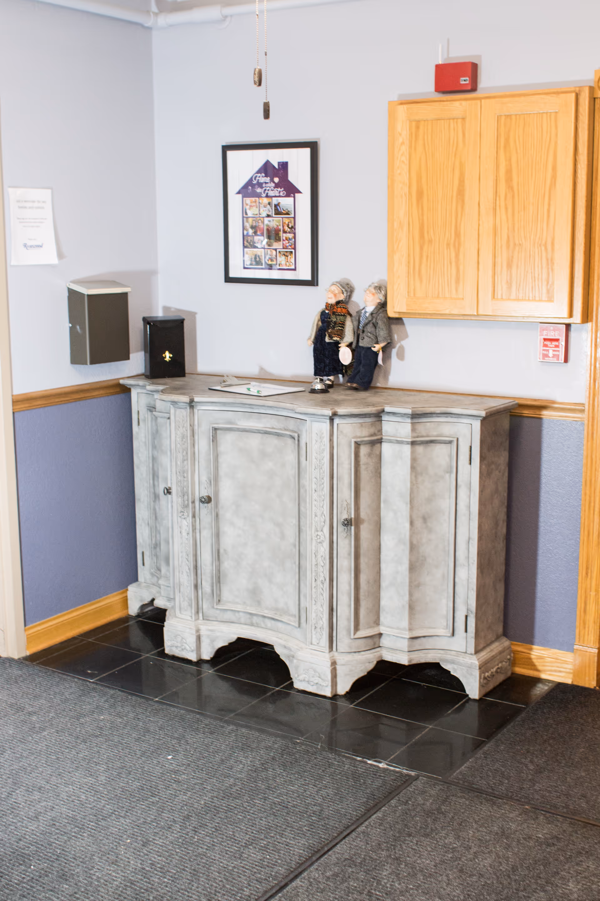 A corner of an interior room with a gray decorative cabinet on a black tiled floor. On top of the cabinet are two small dolls dressed in old-fashioned clothing. Above the cabinet is a wooden wall-mounted cabinet and a framed picture on the wall. The walls are painted light gray with a wooden chair rail and purple lower section. There is a fire alarm and a small black box mounted on the wall.