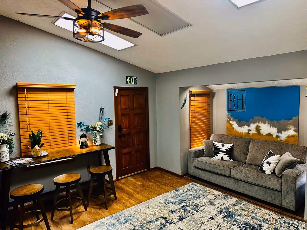 Interior lounge area with couch, bar-height table, stools, art, and window blinds.