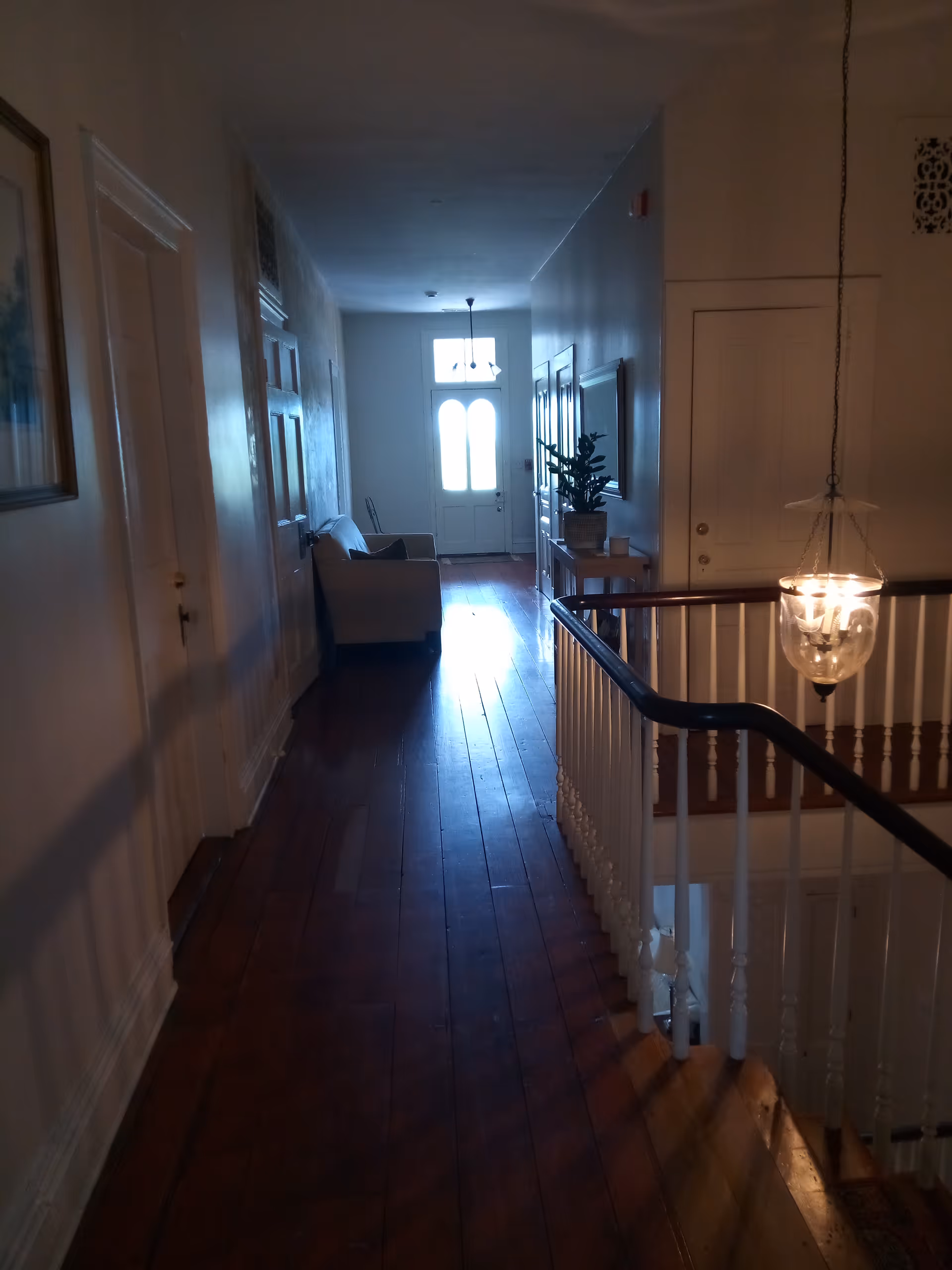 Long interior hallway with wooden floors, railing, chandelier, and a seating area by a front door.