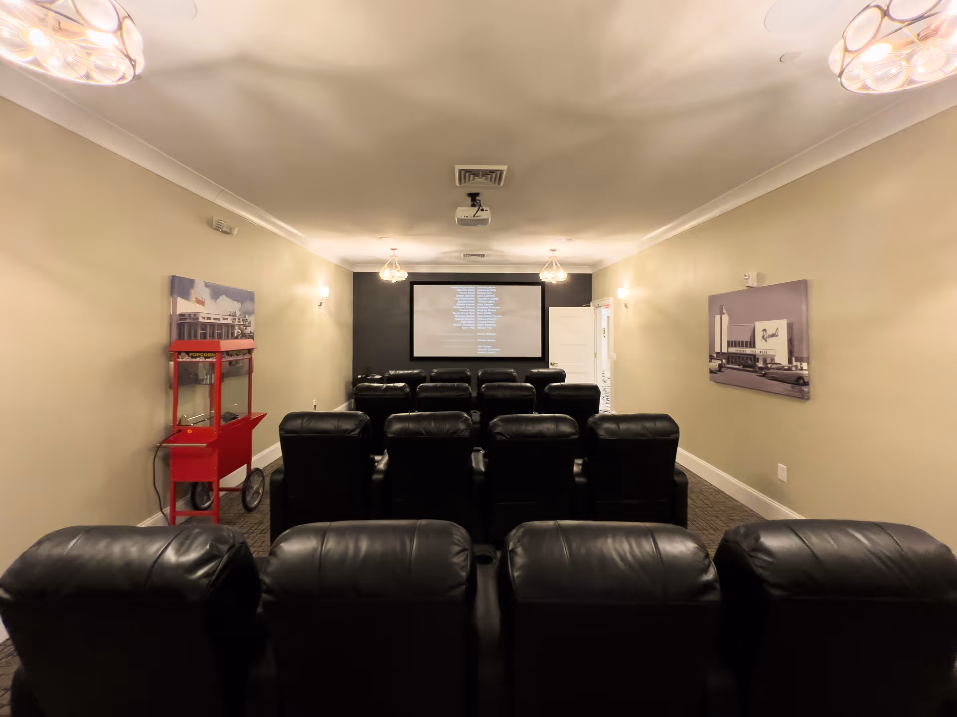 A small movie theater room with several rows of black leather recliner chairs facing a large screen showing movie credits. The room has beige walls, two vintage-style wall lights, a red popcorn machine on the left side, and framed pictures on the walls.